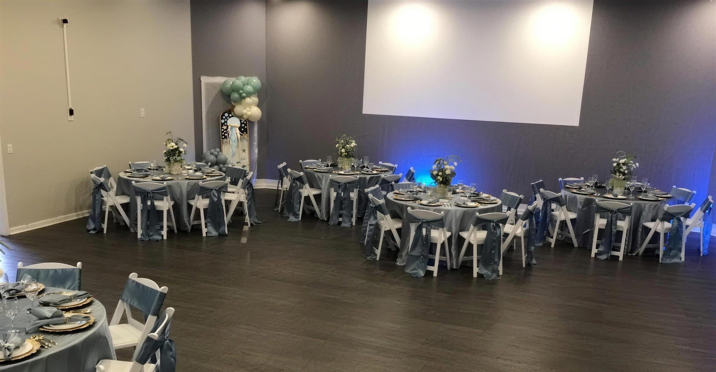 Five round tables in steel blue satin with gold chargers facing a balloon-topped jellyfish backdrop at an under-the-sea birthday at The Cove at St. Johns, St. Johns County FL