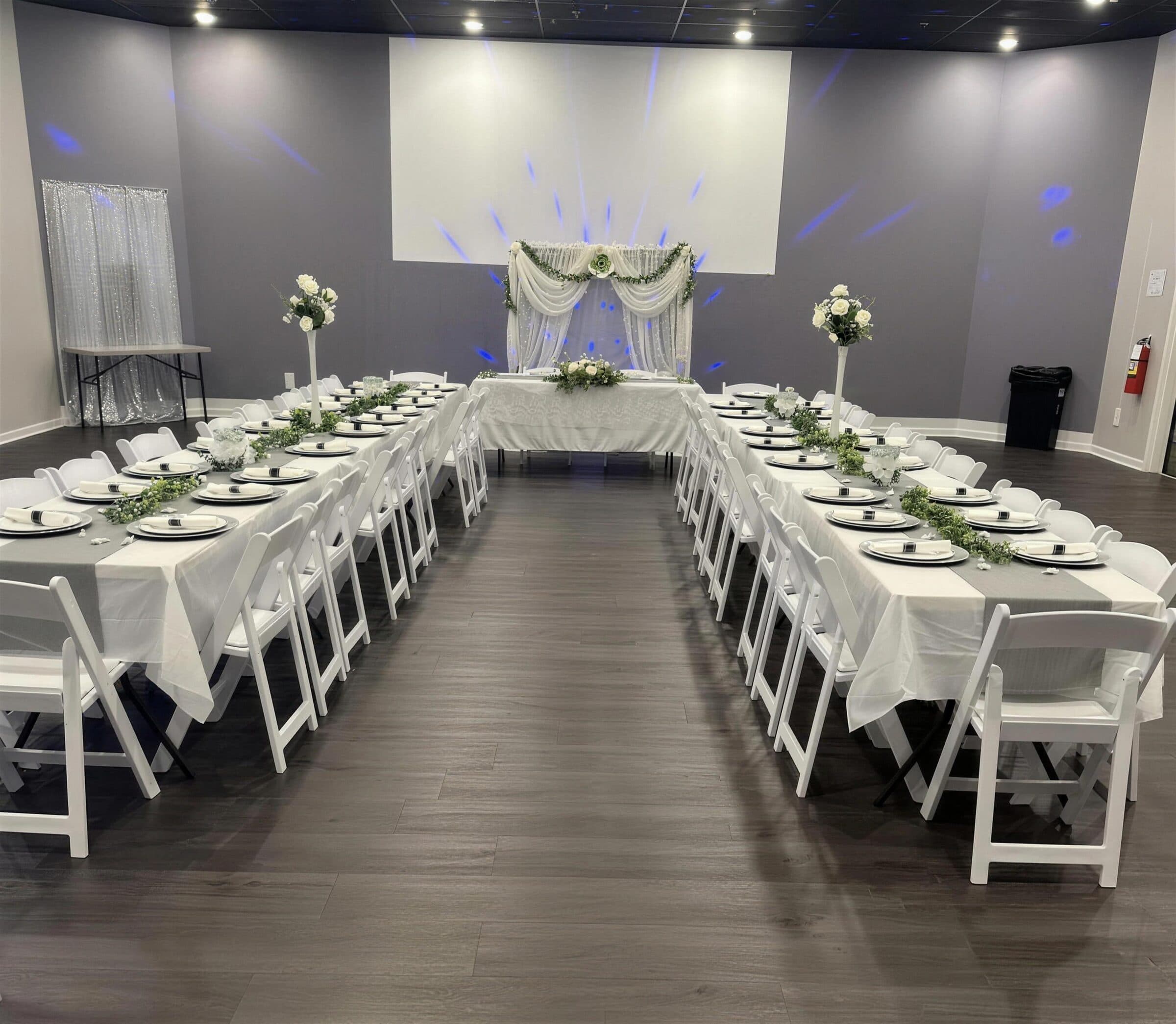U-shaped tables with sage runners, black chargers and tall white rose vases facing a draped sweetheart table with blue uplighting at The Cove at St. Johns, Fruit Cove FL